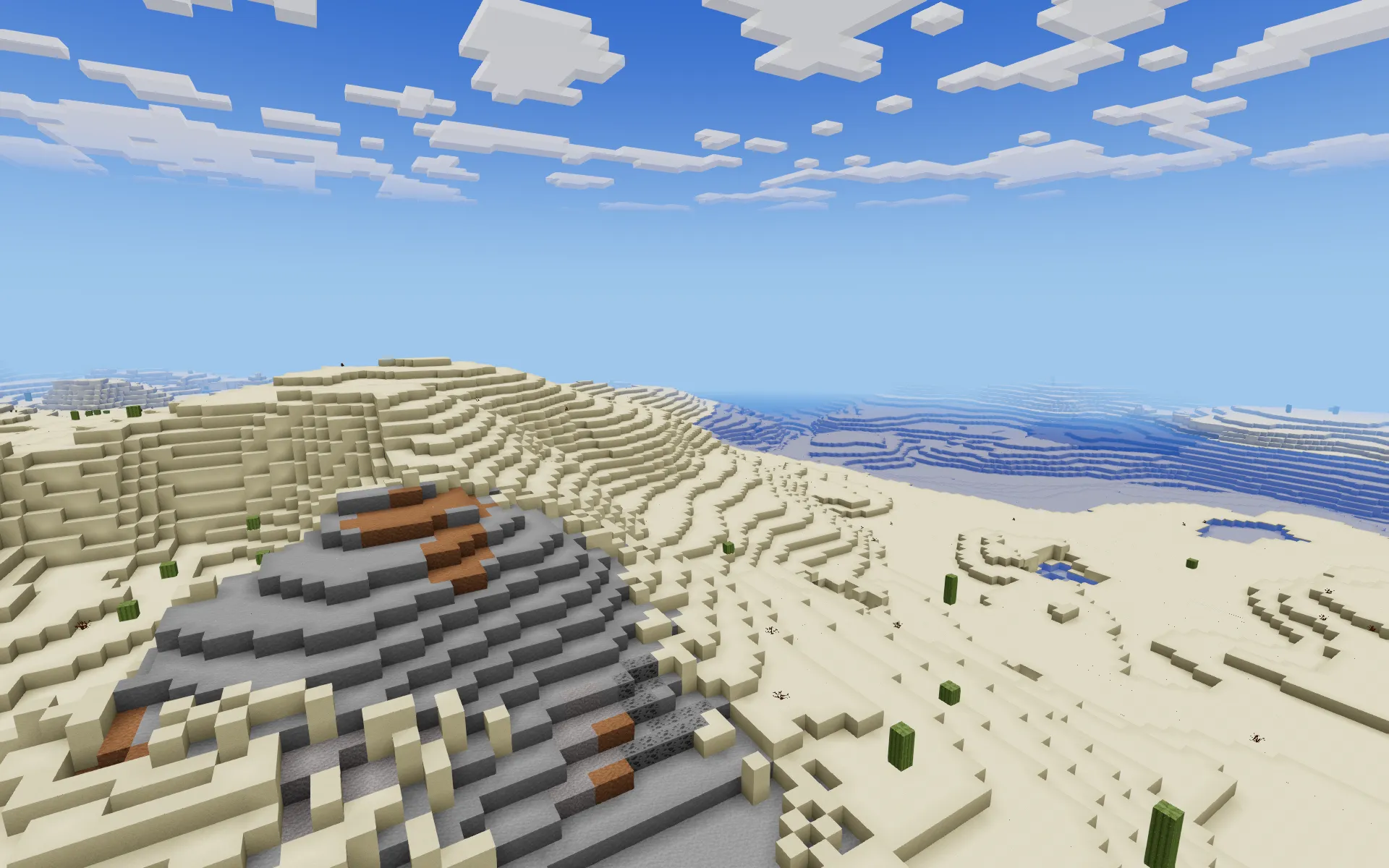 Pale sand dunes around a grey stone outcrop streaked with ore, with scattered cacti and the sea on the horizon.