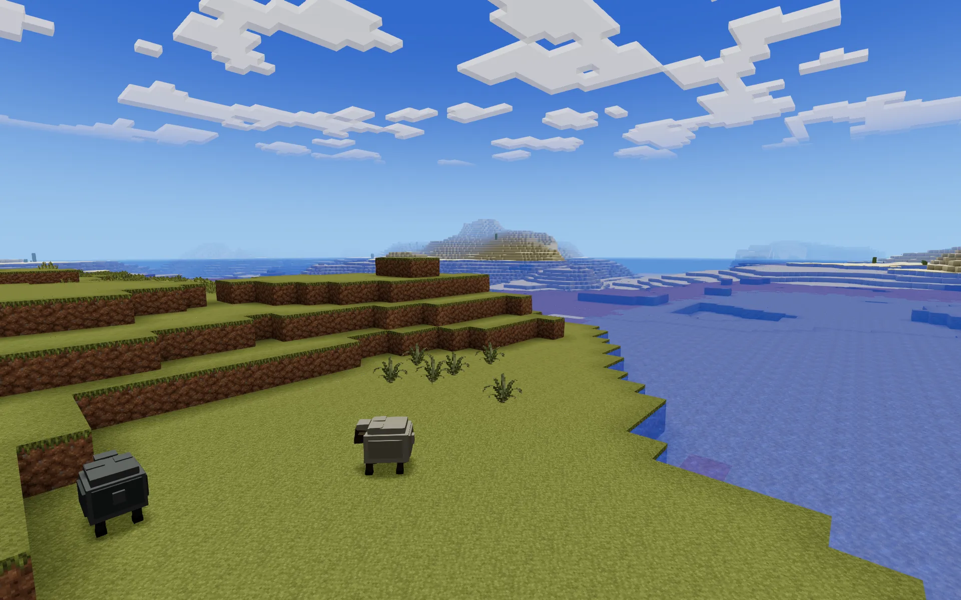 Two sheep on a grassy shore beside a calm blue lake, with stepped dirt terraces and ferns behind them.