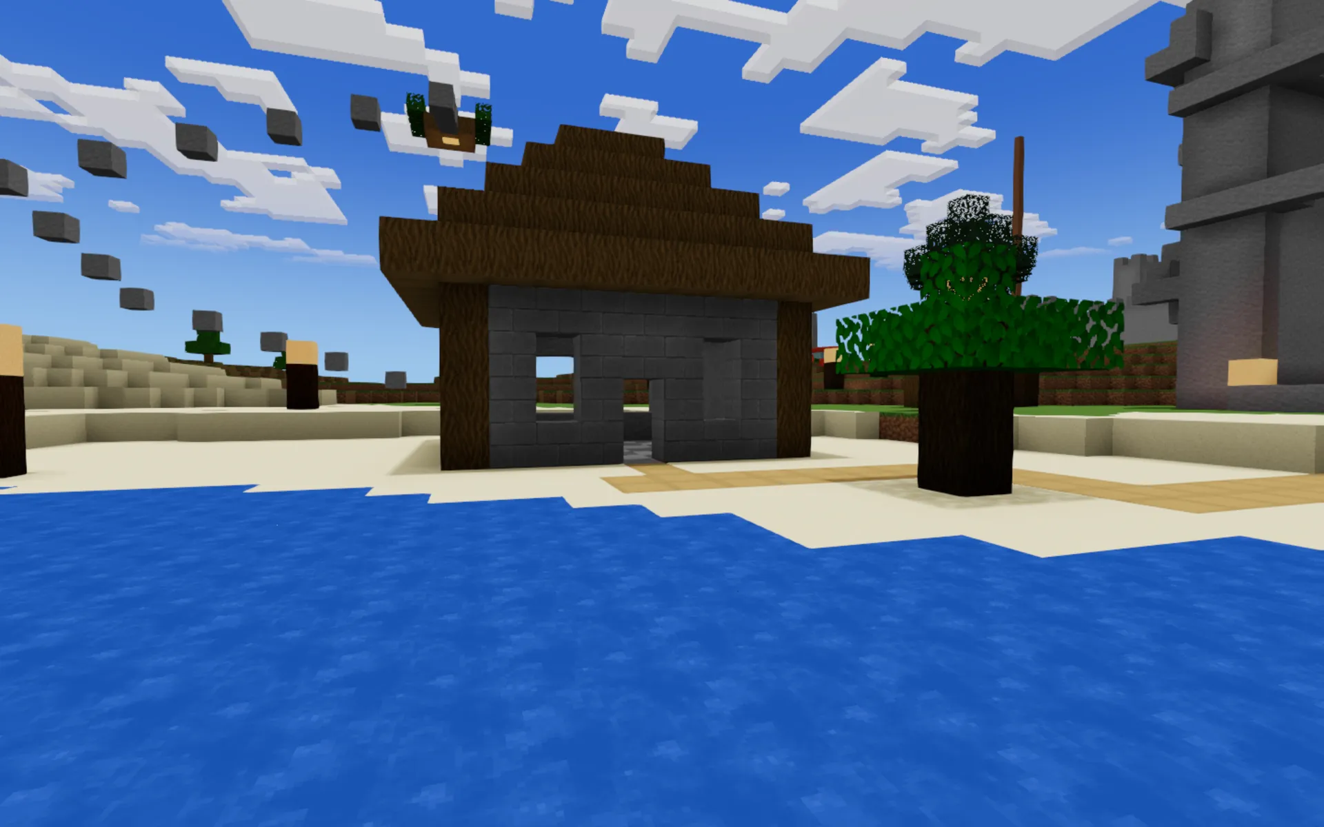 A player-built stone house with a wooden roof standing on the sand beside blue water.