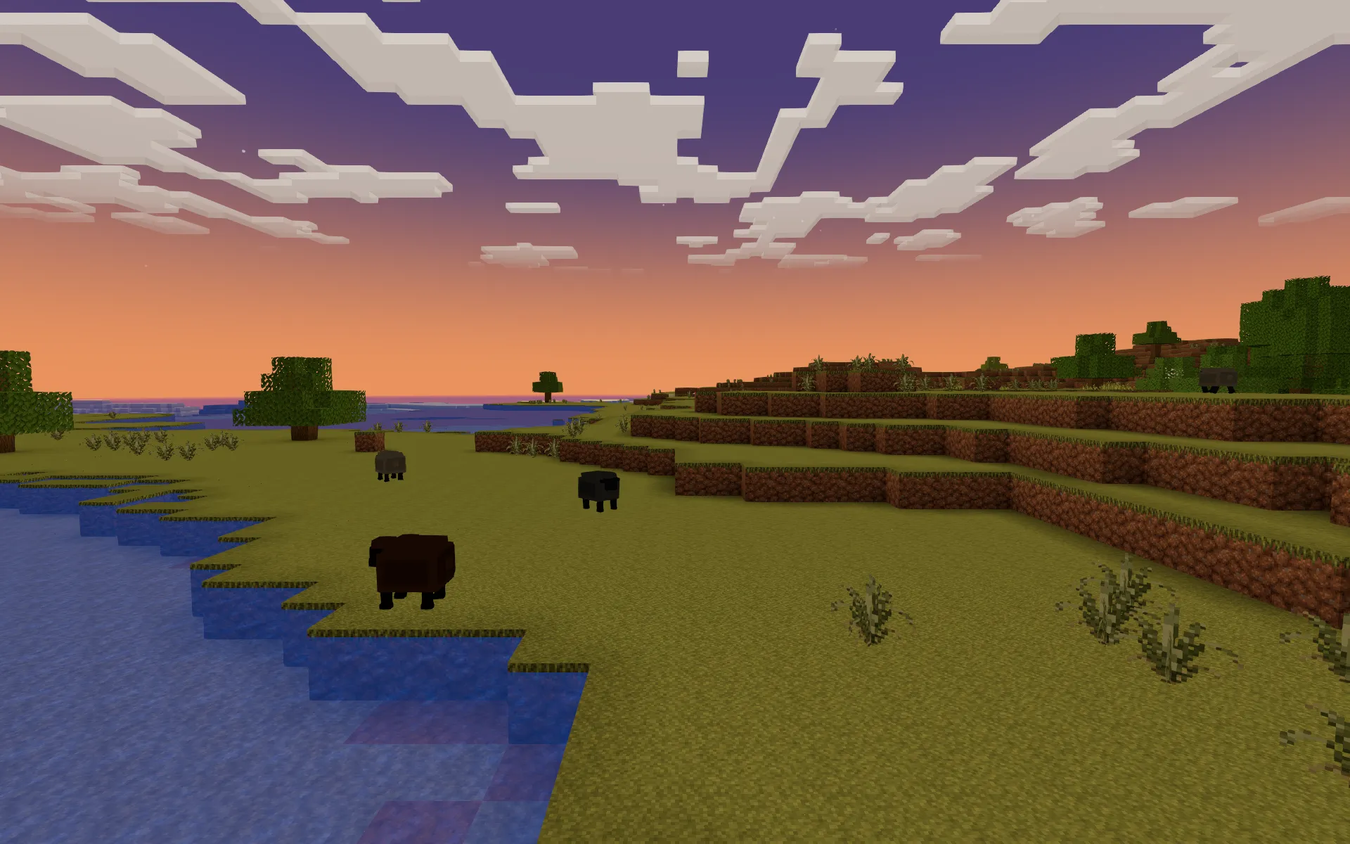 A small flock of sheep grazing on a grassy lakeshore beneath an orange and violet sunset sky with blocky clouds.